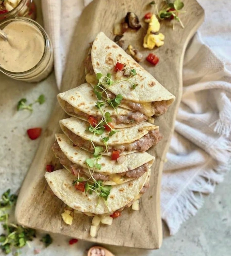 Six folded beef and bean quesadillas topped with microgreens and diced red pepper, served on a wooden serving board with a small jar of sauce and a white cloth in the background.