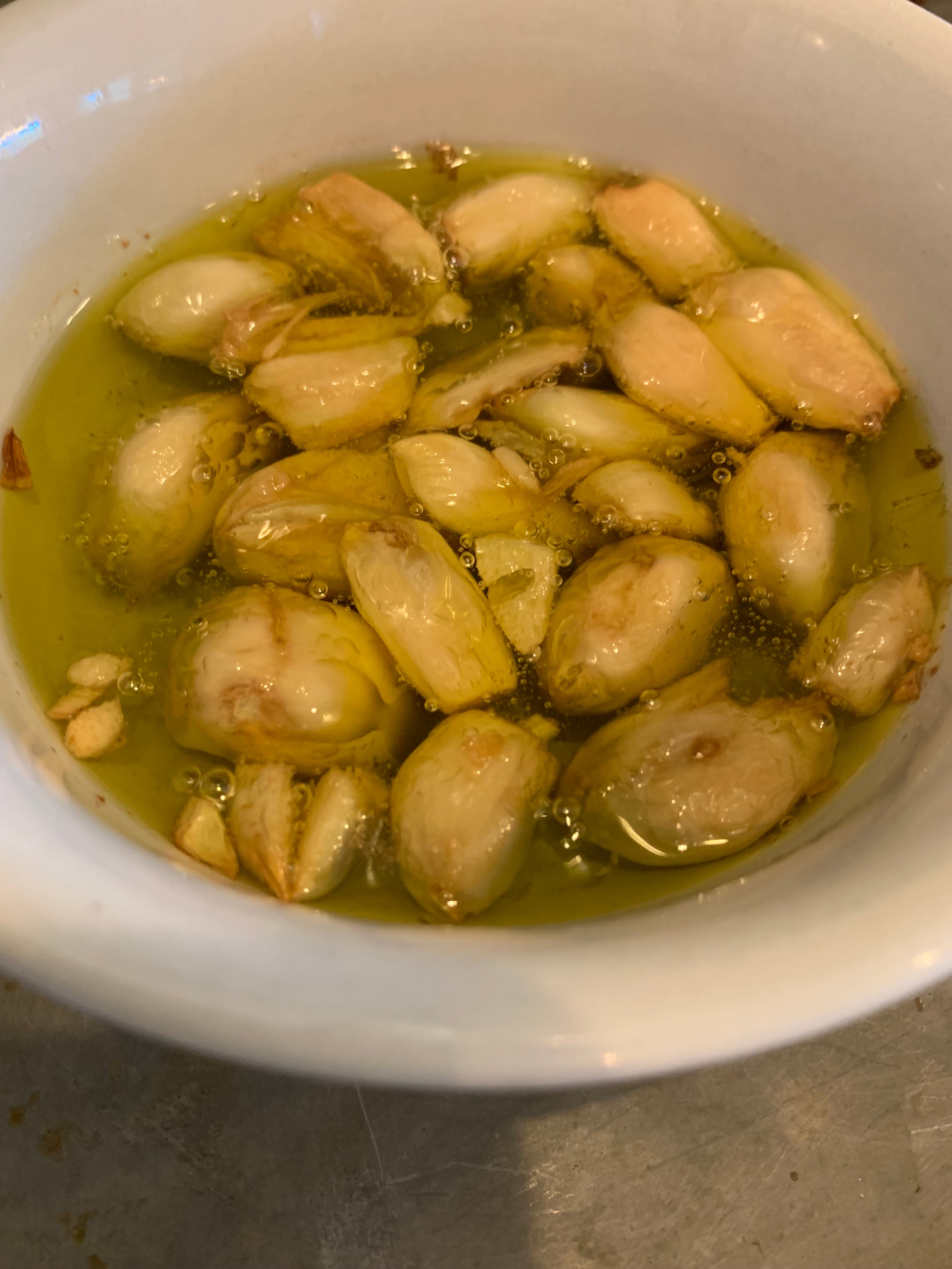 Crispy fried garlic cloves in a light olive oil sauce, served in a white ceramic bowl on a gray surface.