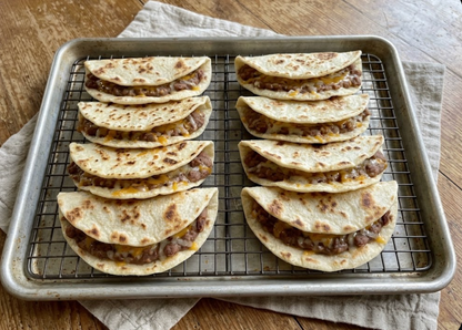 Four stacks of soft corn tortilla tacos filled with seasoned ground meat and melted cheese, arranged on a wire cooling rack set inside a metal baking sheet on a wooden table.